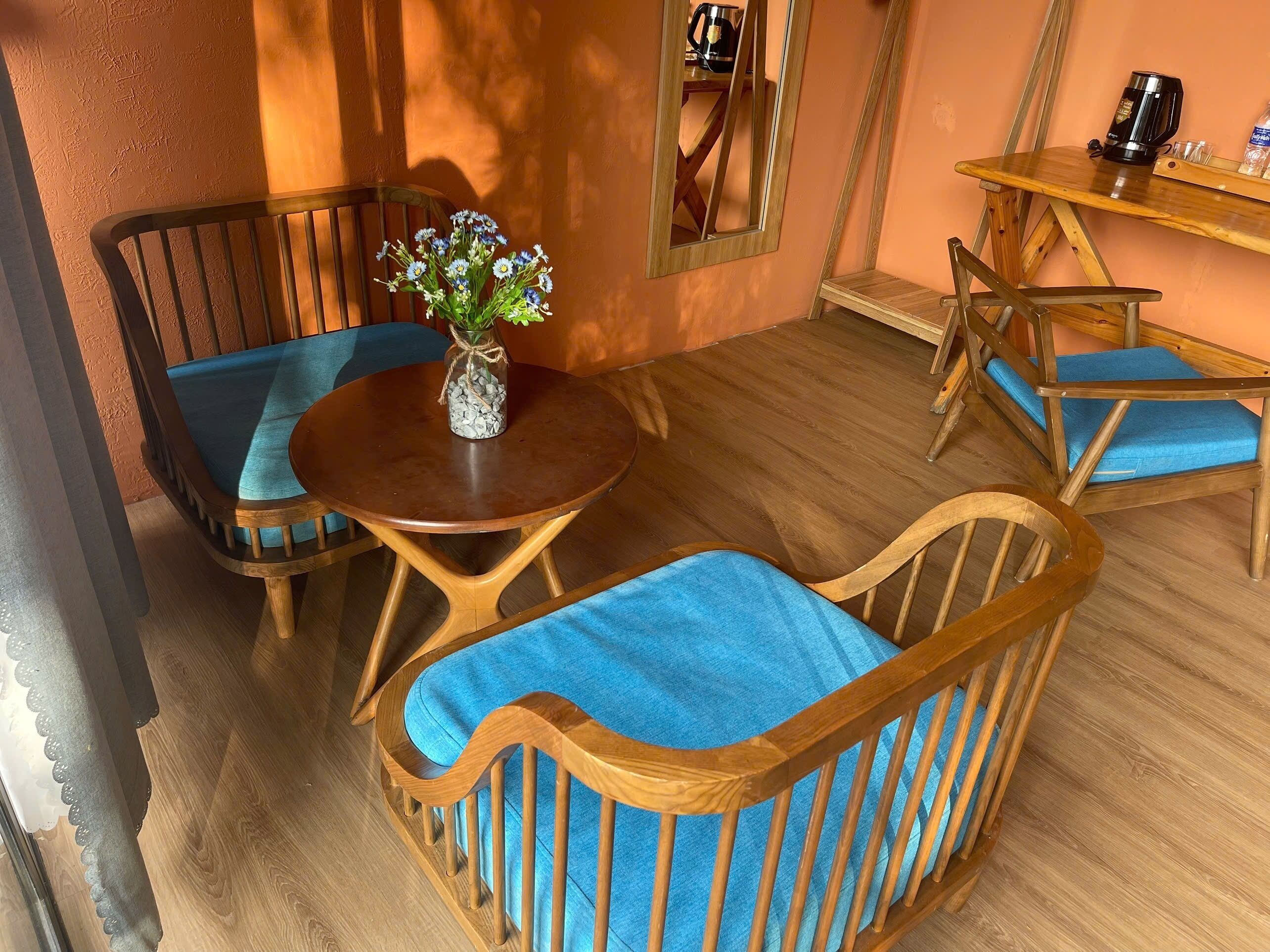 A room with a round wooden table, two wooden chairs with blue cushions, a vase with flowers, and a coffee machine on a wooden shelf.