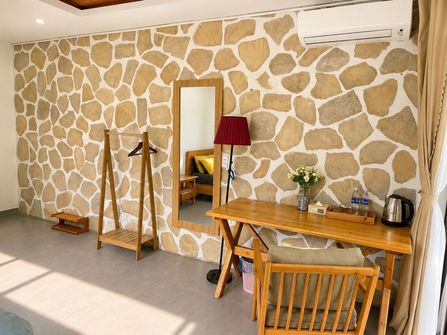 A room with a stone wall, a wooden desk, a chair, a mirror, a red lamp, and a hanging coat rack.