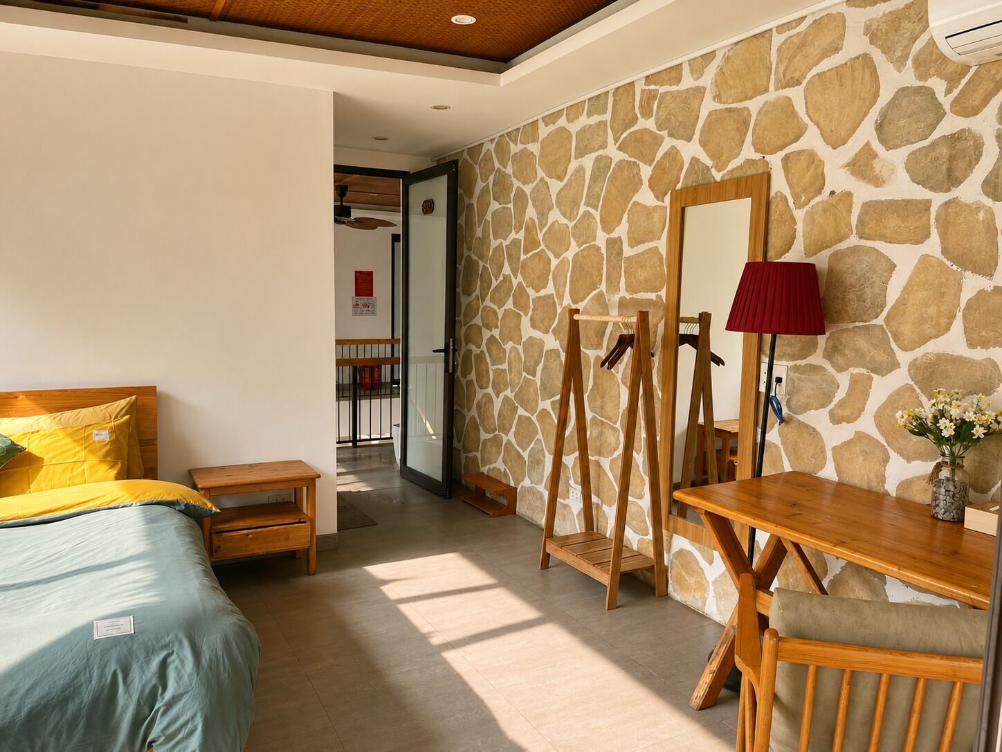 A hotel room with a bed, a wooden desk, a mirror, and a stone wall.