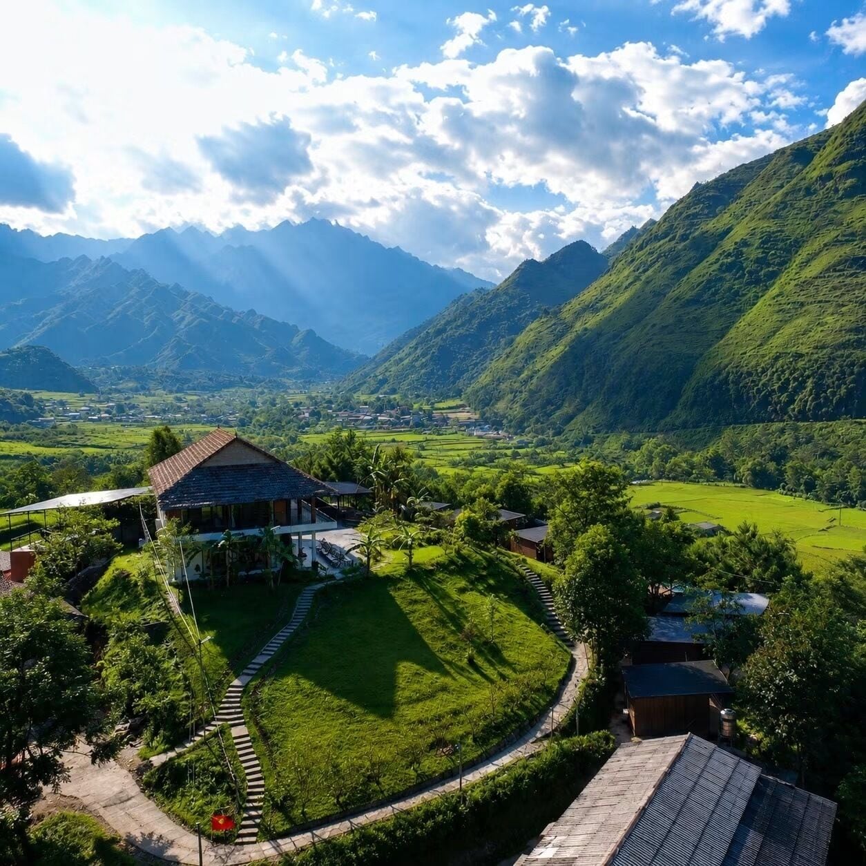 A rural landscape with a house surrounded by lush greenery and mountains in the background.