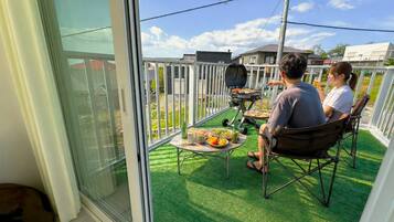 Un balcón con una parrilla, una mesa con comida y vistas del barrio.
