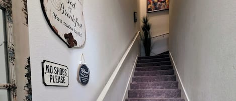 A staircase with a 'NO SHOES PLEASE' sign, a 'Welcome' sign, and a decorative wall piece.