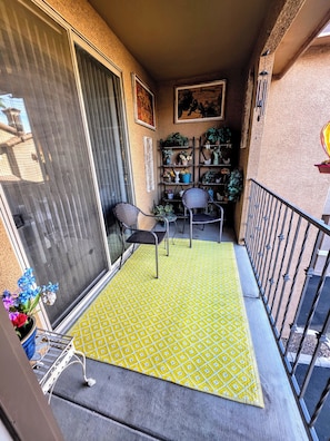 Un balcon avec un tapis jaune à motifs, deux chaises en osier, une plante en pot et une rambarde en métal.