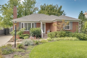 A two-story brick house with a front porch, a chimney, and a well-maintained front yard with various plants and a lawn. - Near Universities & Hospitals! SLC Home w/ Yard (Salt Lake City)