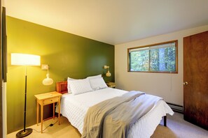 Ein Schlafzimmer mit Bett, Nachttisch, Lampe und einem Fenster mit Blick auf Bäume.