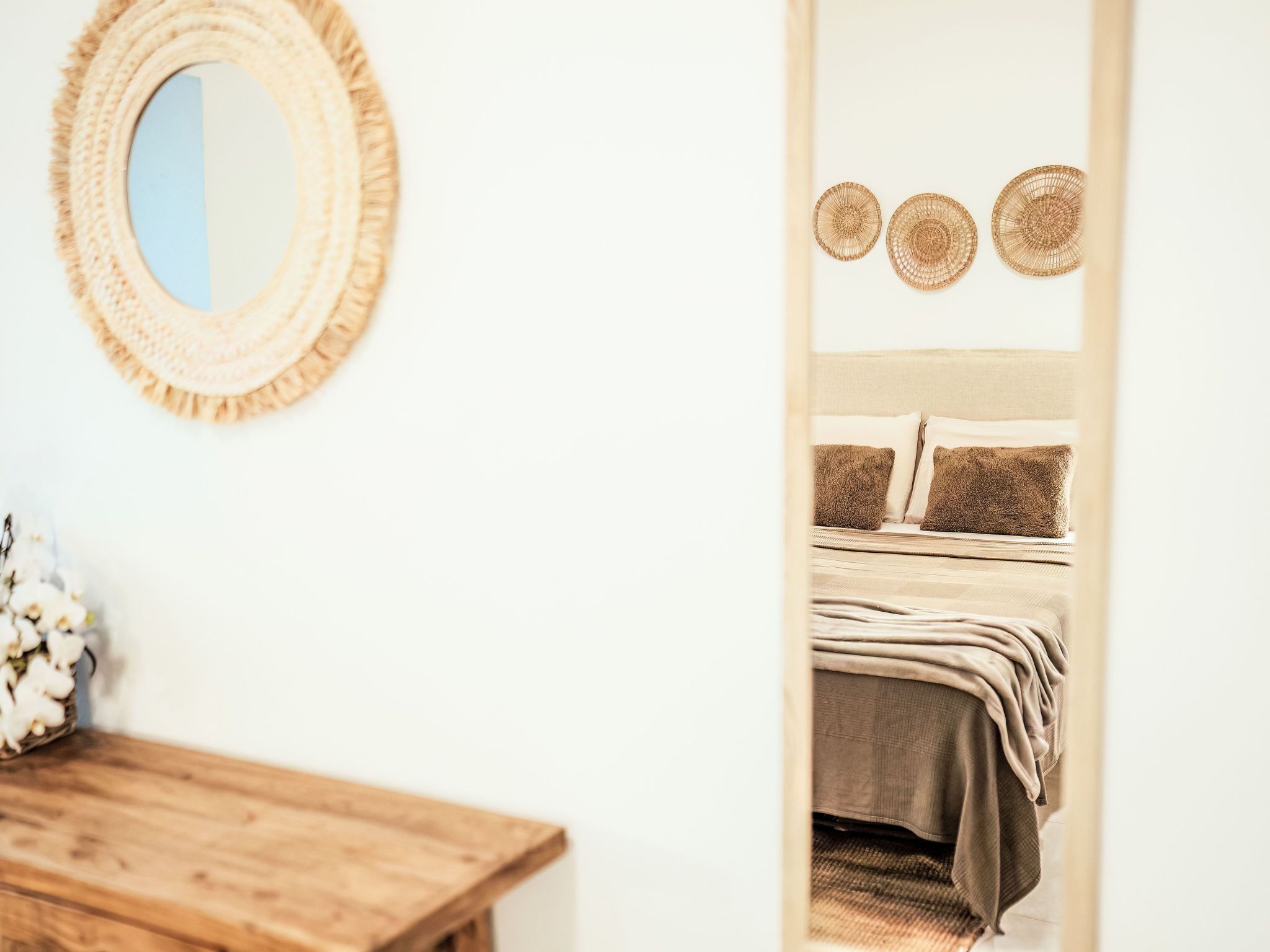 A bedroom with a bed, pillows, a mirror, and a wooden table with a vase of flowers.