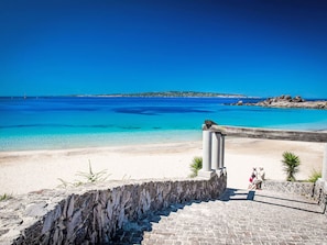 Vistes costaneres amb un mar blau clar, una platja sorrenca i un camí de pedra que condueix a una estructura amb columnes.