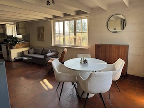 A dining area with a round table and chairs, a sofa, a window with a view of trees, and a kitchen in the background.