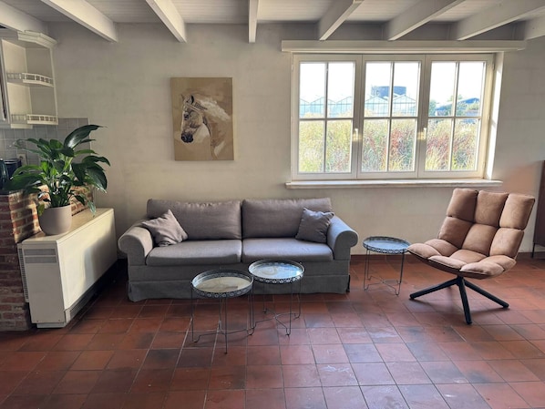A living room with a grey sofa, two small round tables, a brown armchair, a potted plant, a painting of a horse, and a large window with a view.