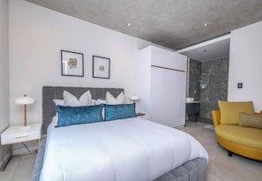 Ein modernes Schlafzimmer mit einem großen Bett, Nachttischen, einem Sessel und einem durch eine offene Tür einsehbaren Badezimmer.