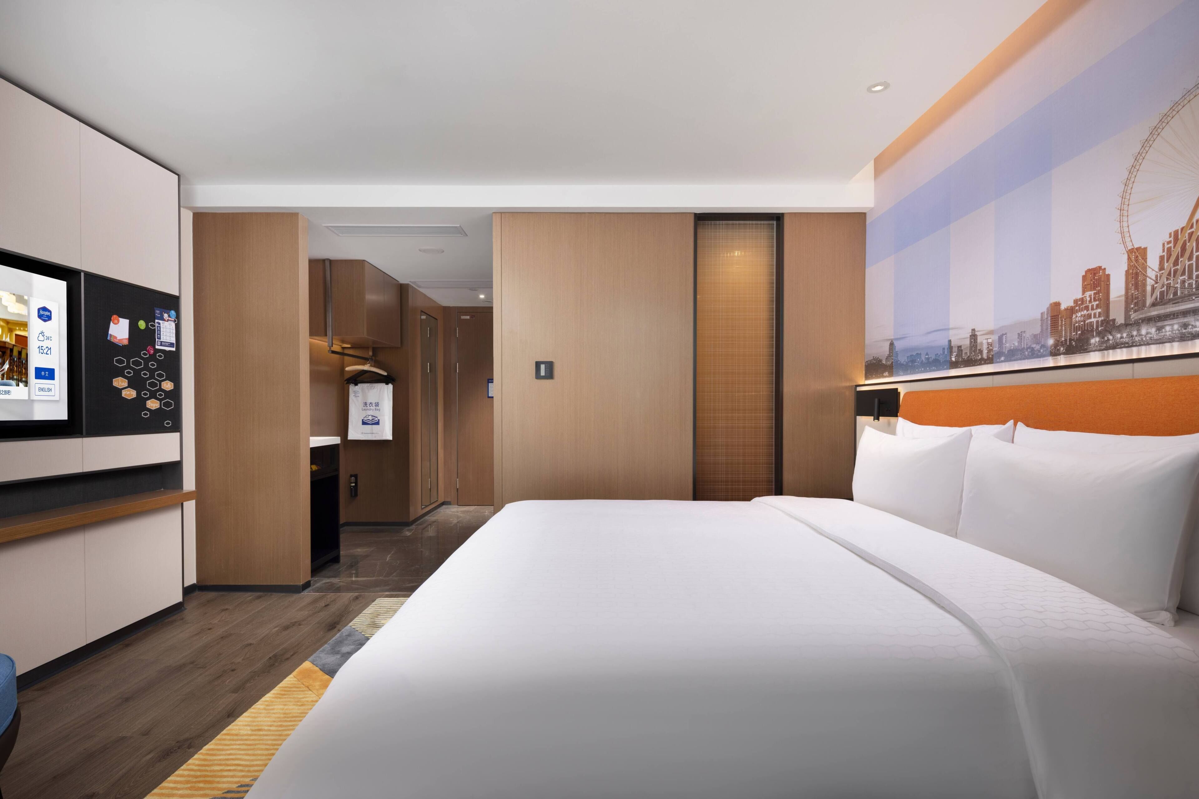 Ein Hotelzimmer mit einem großen Bett, einem Fernseher, einem Kleiderschrank und Blick auf die Stadt.