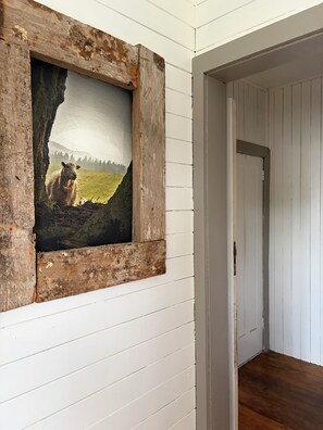 A rustic wall with a framed painting of a landscape, a wooden door, and a hallway with white paneling.