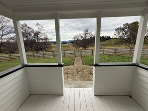A covered porch with white siding and black trim, leading to a grassy area with a wooden fence and trees.