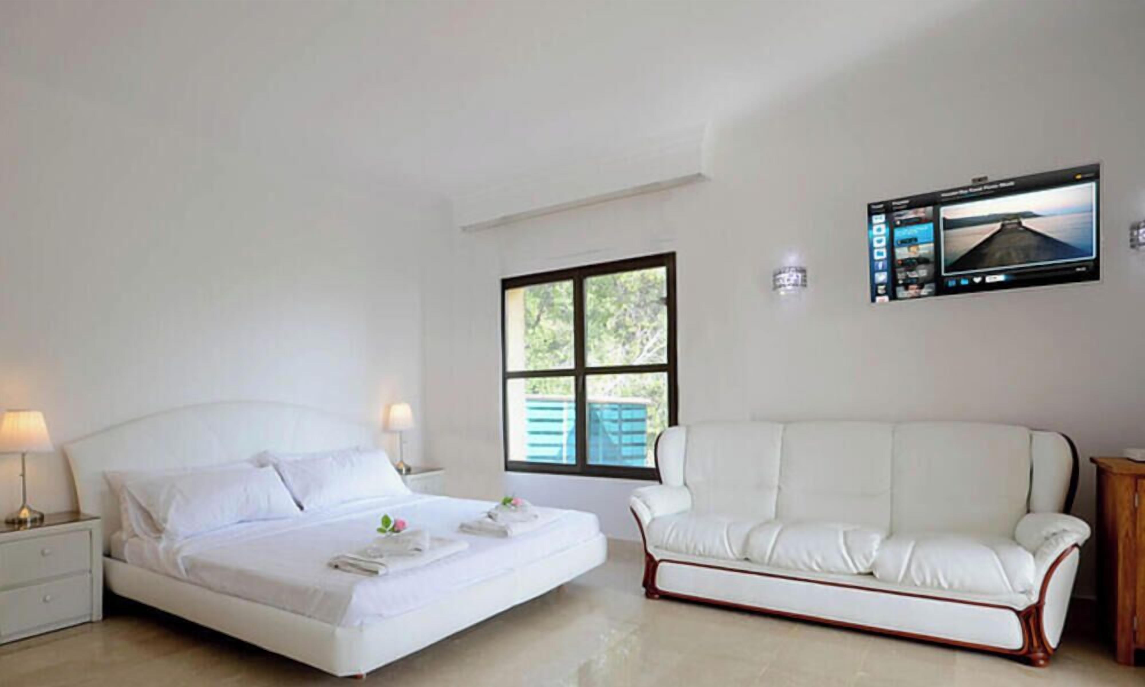 Une chambre d’hôtel avec un lit, un canapé blanc, un téléviseur fixé au mur et une fenêtre donnant sur un paysage verdoyant.
