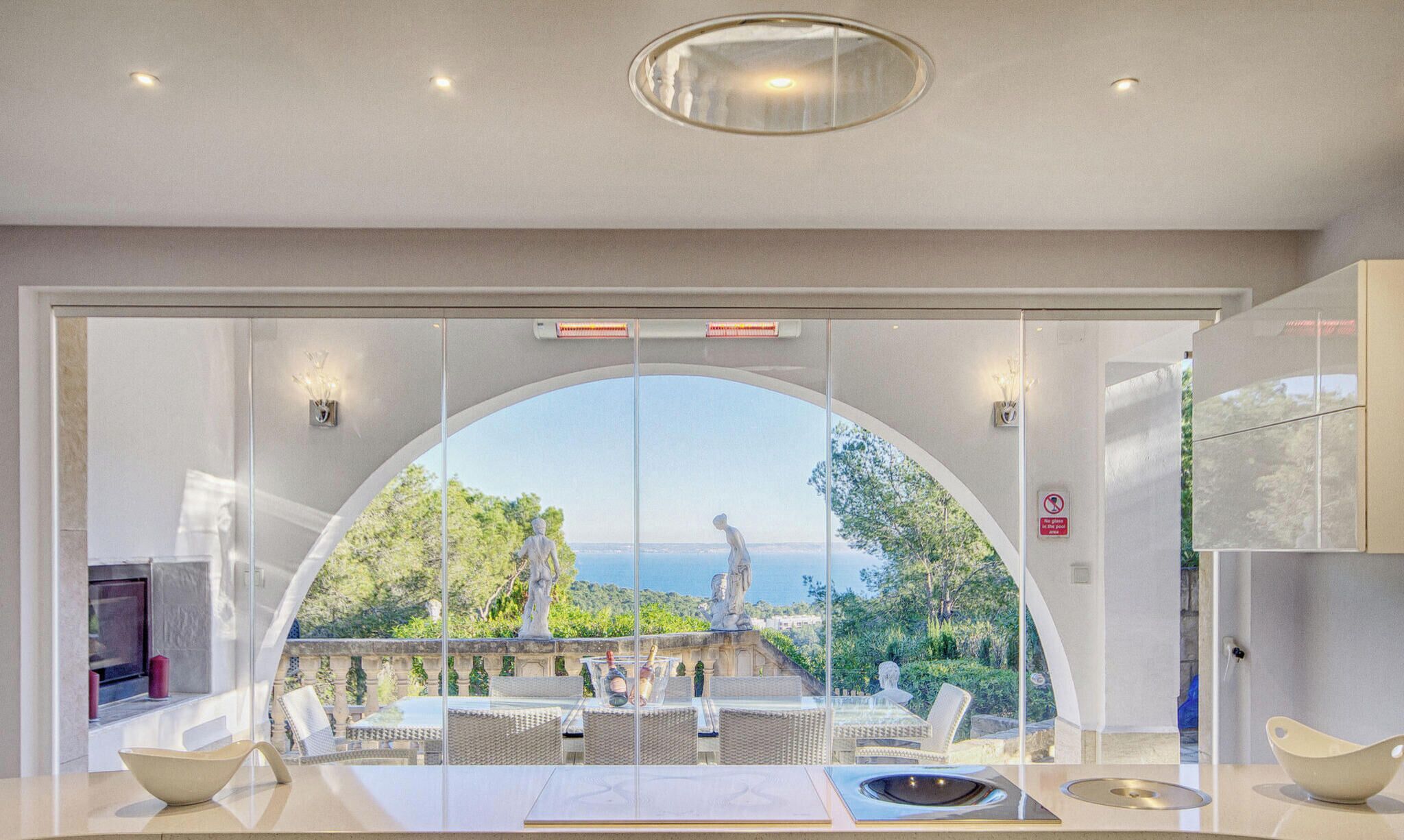 Un intérieur moderne avec une grande porte cintrée en verre donnant sur une terrasse extérieure offrant une vue sur la mer et ornée de statues.
