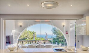 Ein modernes Interieur mit einer großen, bogenförmigen Glasdoor, die auf eine Außenterrasse mit Meerblick und Statuen führt.