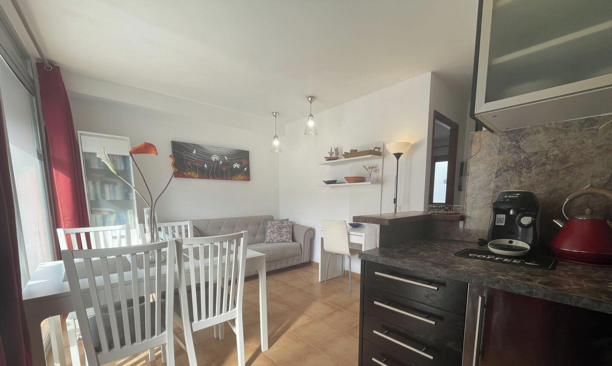 Eine moderne Küche mit Kaffeemaschine, ein Essbereich mit weißem Tisch und Stühlen sowie ein Wohnzimmer mit Sofa und einem Bild an der Wand.