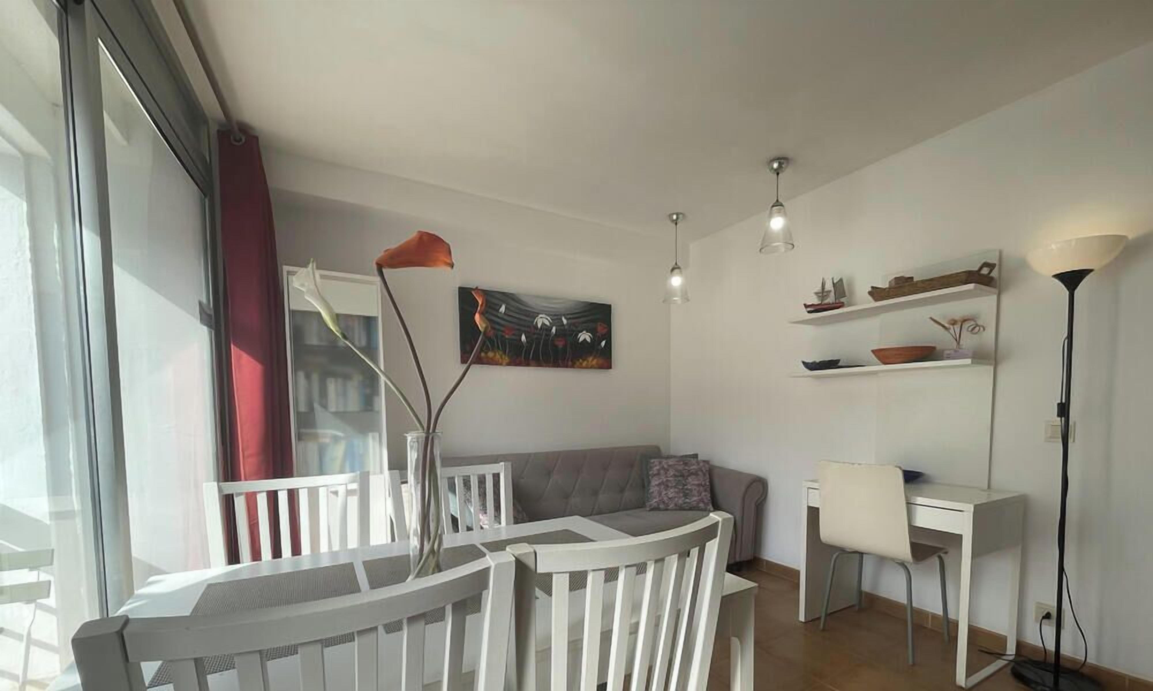 Een moderne woonkamer met een witte bank, een eettafel en een wit bureau. Er hangt een schilderij aan de muur en er staat een vloerlamp.