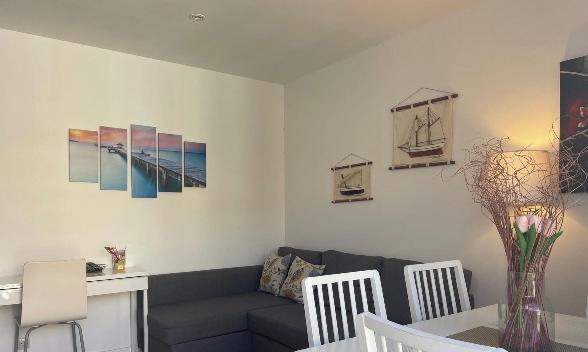 Un salon moderne avec un canapé gris, une table à manger entourée de chaises blanches et des œuvres d’art accrochées aux murs.