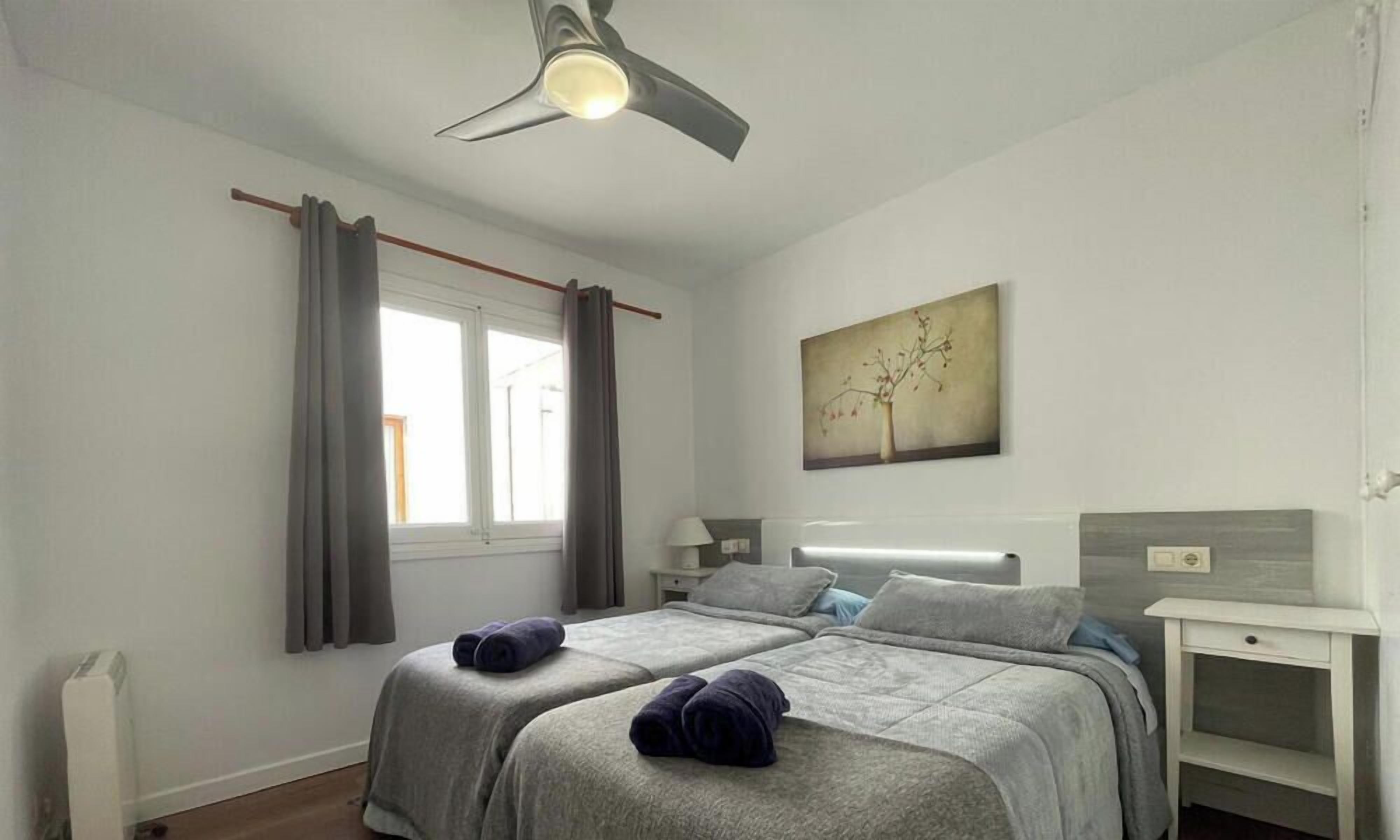 Un dormitorio con dos camas, un ventilador de techo, una ventana con cortinas, una mesita de noche y una obra de arte colgada en la pared.