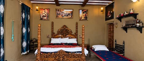 A traditional four-poster bed with ornate carvings, a single bed with a patterned bedspread, and a shelf with kitchenware.