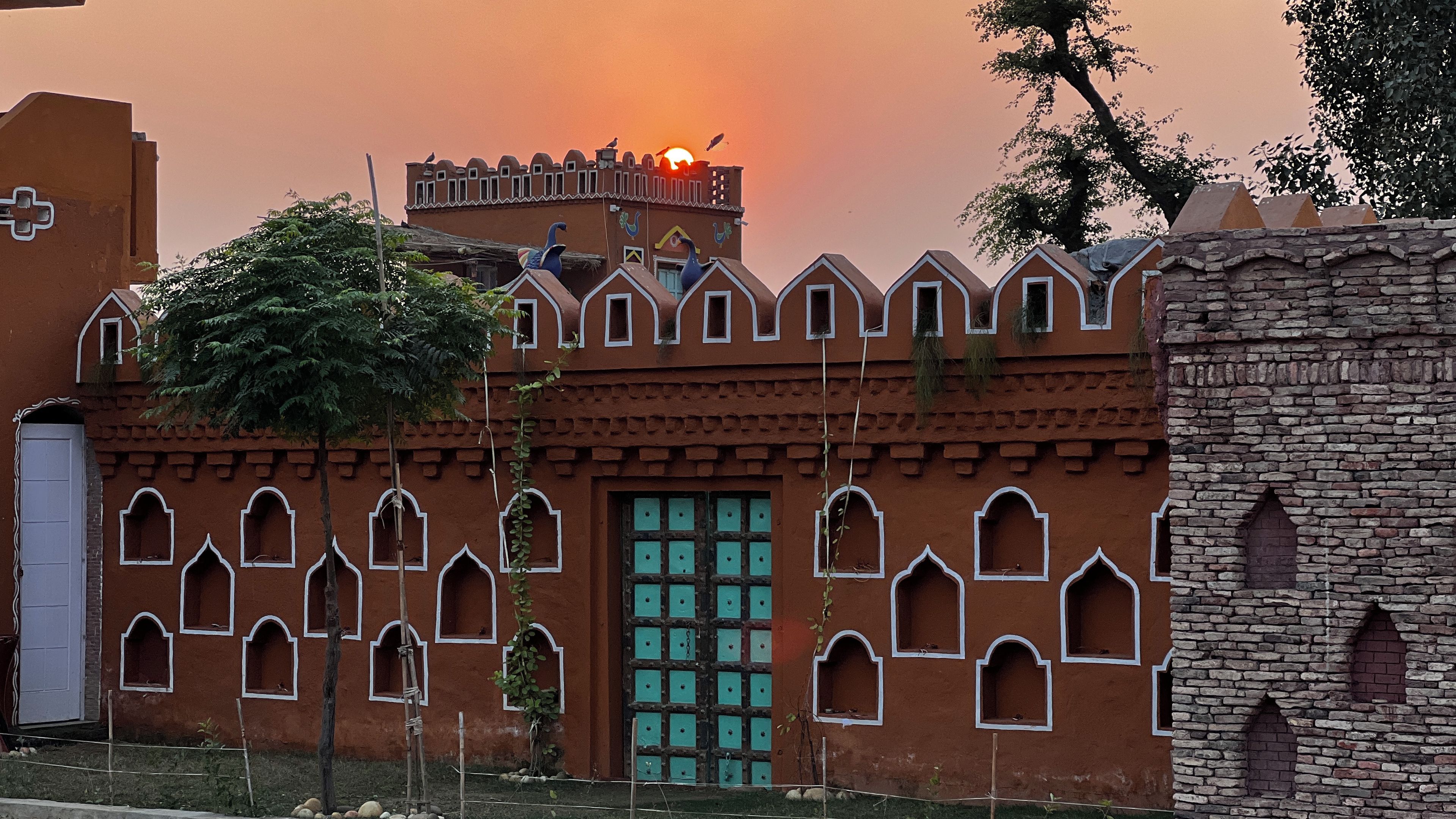 Un edificio tradicional con ventanas arqueadas y una puerta central, rodeado por un muro bajo y un árbol.