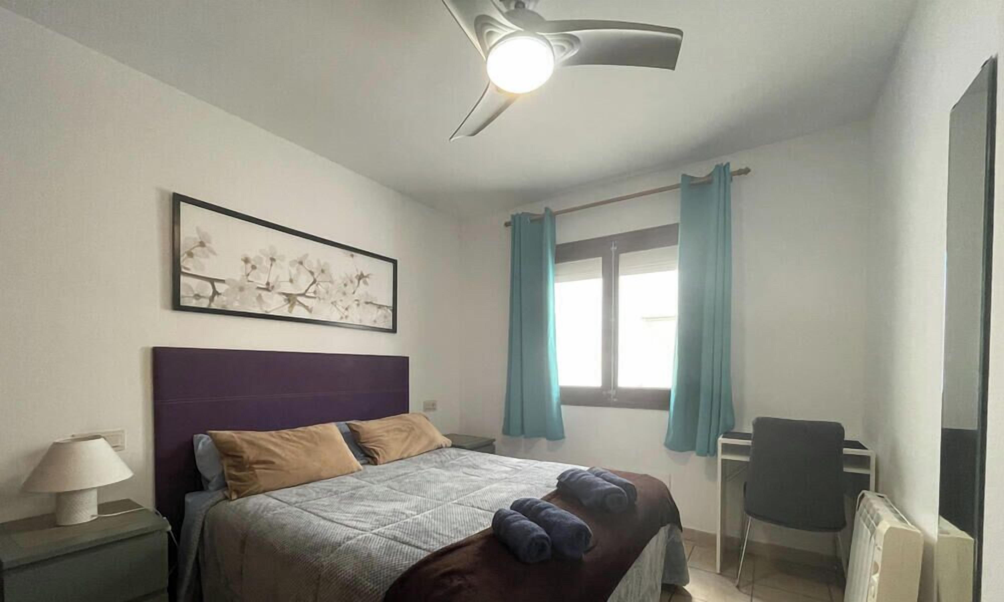 Un dormitori amb llit, escriptori, cadira, ventilador de sostre i finestra amb cortines.
