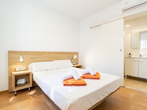 Ein Hotelzimmer mit hölzernem Kopfteil, einem Bett mit weißen Laken, zwei zusammengerollten Handtüchern, einem Nachttisch mit Lampe, einer kleinen Ablage und einem Badezimmer mit Spiegel und Schrank.