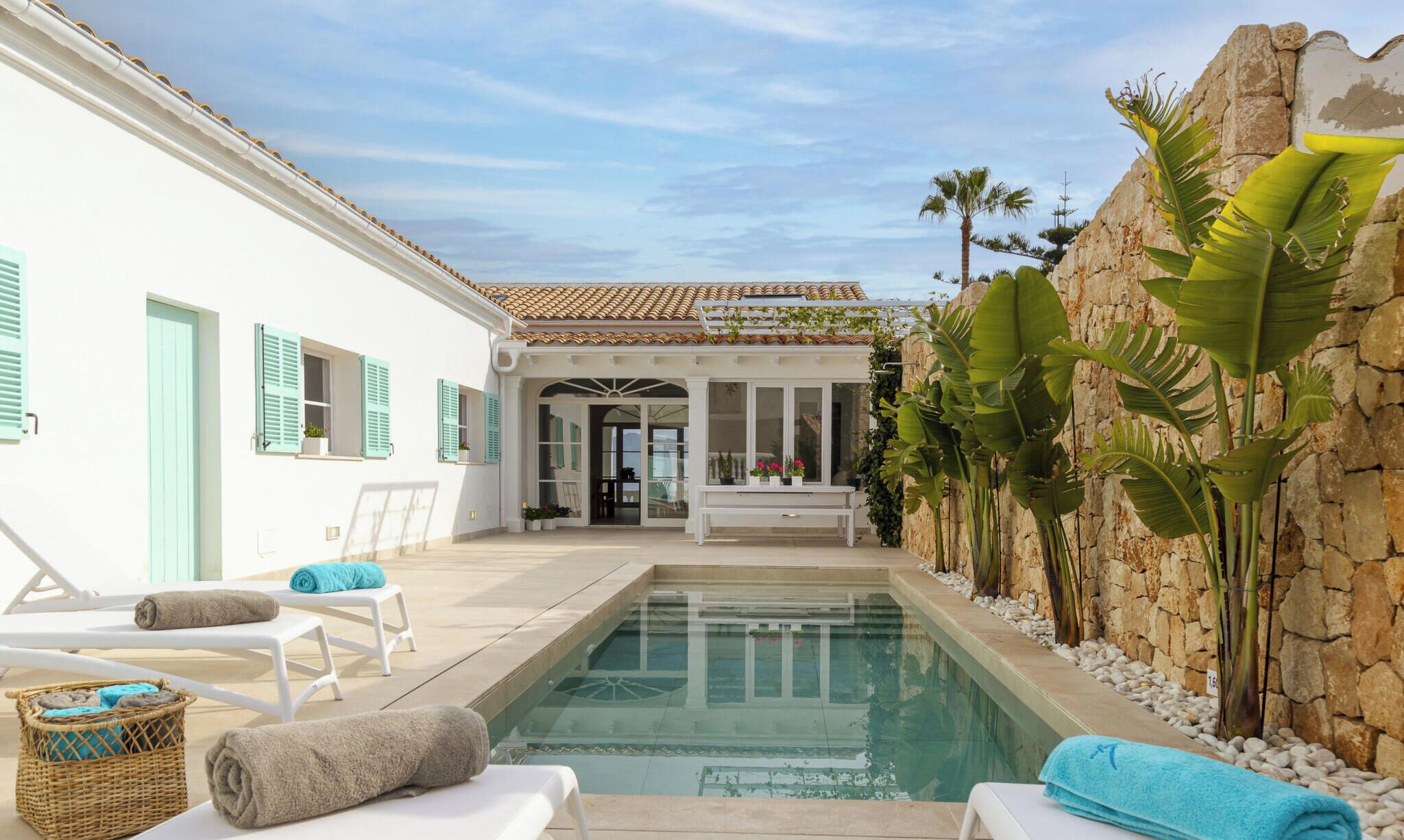 A pool area with lounge chairs, a bench, and a stone wall with tropical plants.