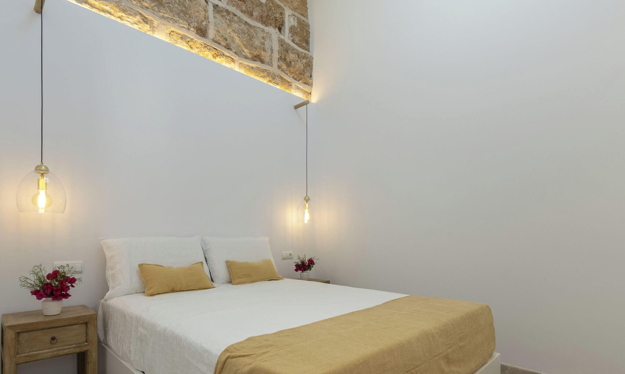 A bed with white linens, two pillows, and a beige throw. There are two bedside tables with lamps and flowers. The room has plain white walls and a stone accent wall.
