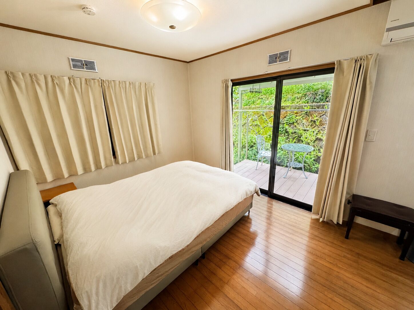 A bedroom with a bed, a bench, a window with curtains, and a view of greenery outside.