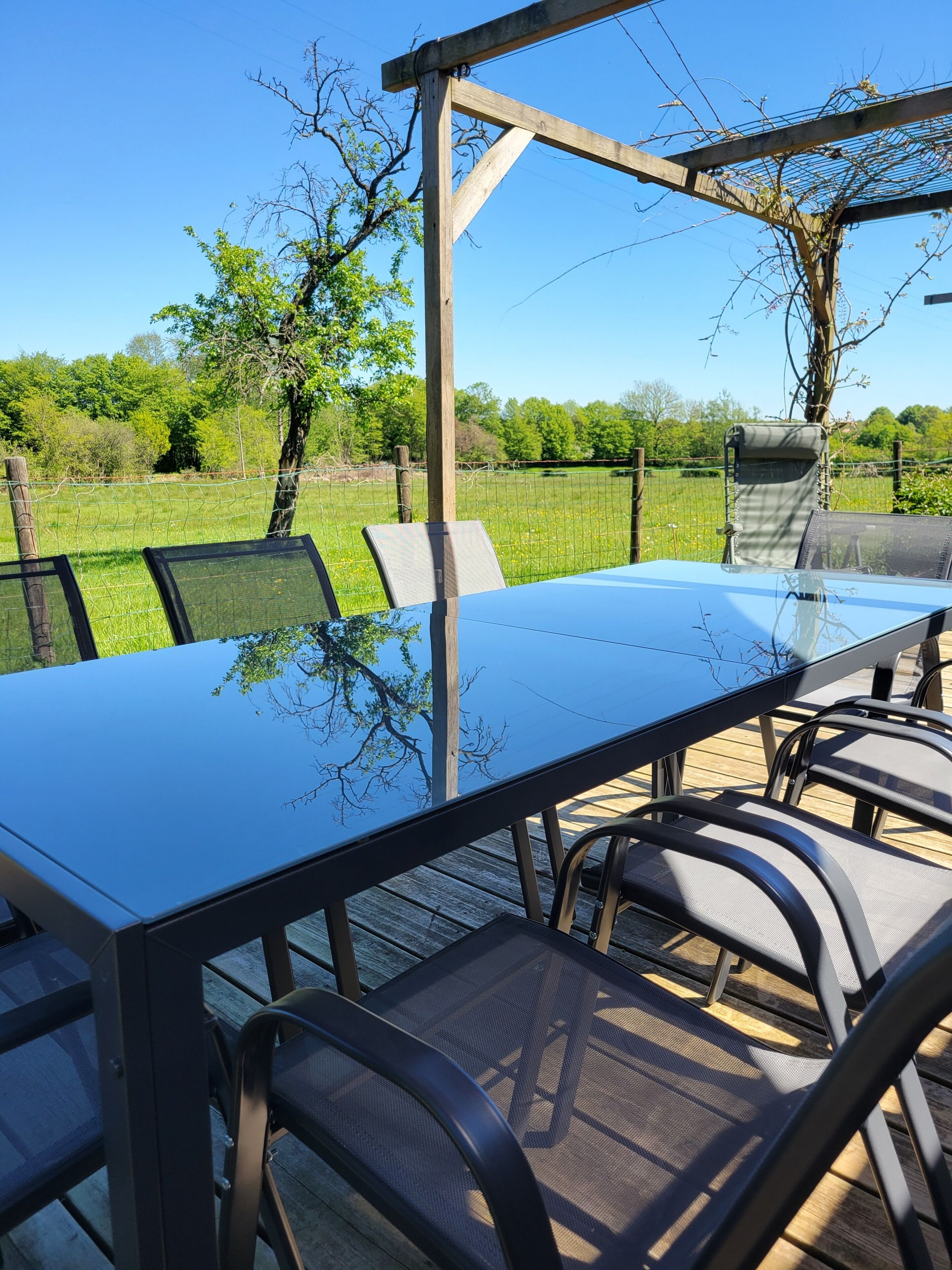Un área de comedor al aire libre con una mesa de vidrio y sillas de metal, rodeada por una pérgola de madera y una cerca de alambre.