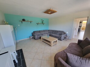 A living room with a sectional sofa, a coffee table, and a TV. There is a kitchen area in the background. - Large vacation rental for 10 people with a sauna, garden, and fireplace (Anor)