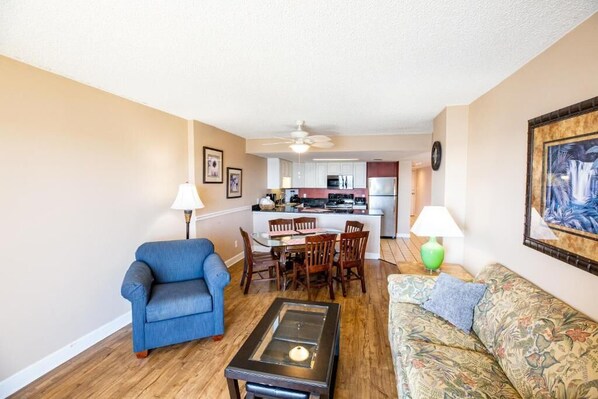 A living room with a blue armchair, a patterned sofa, a coffee table, and a dining area with a table and chairs. - Atalaya Towers - 3 Bedroom (Murrells Inlet)