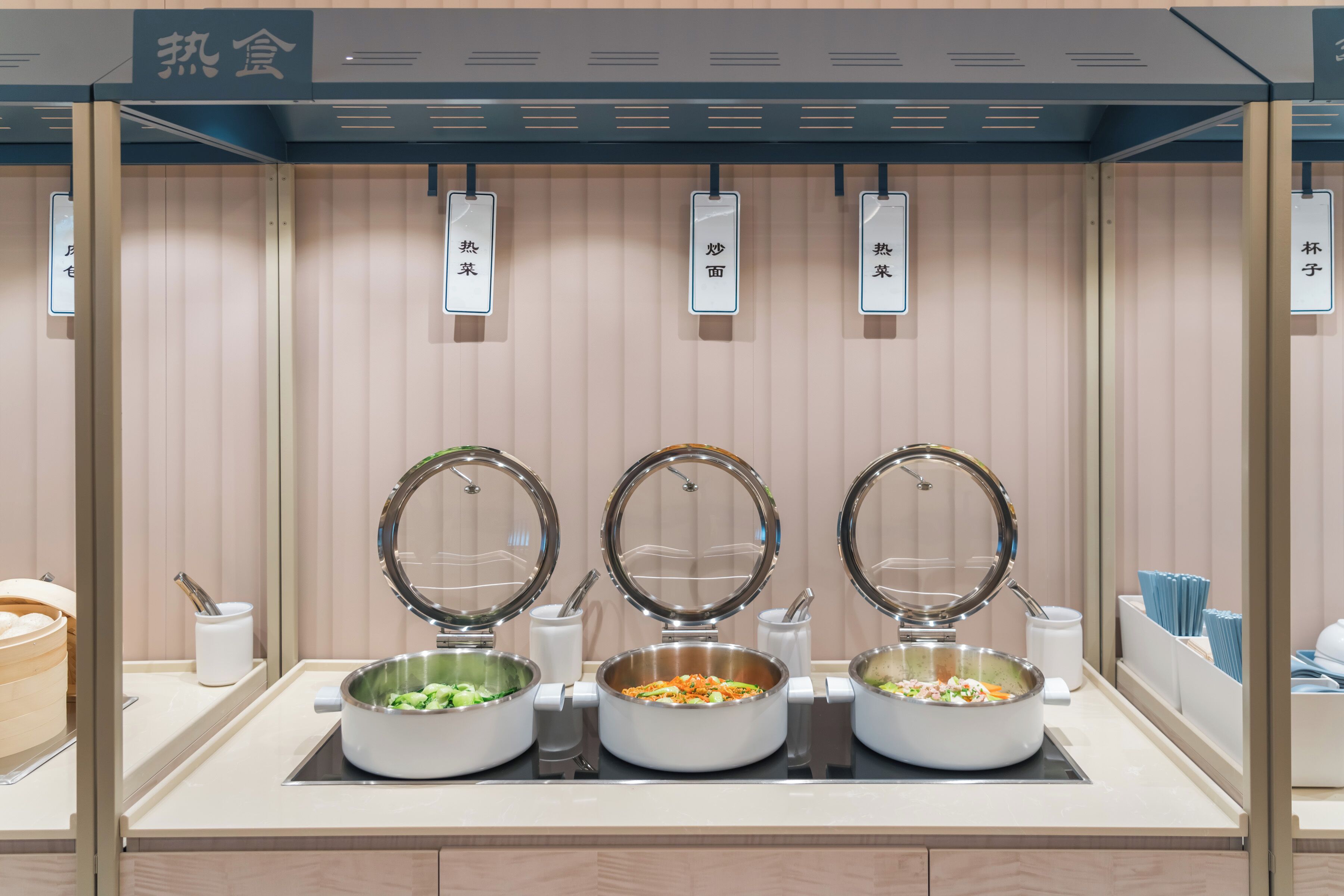 Un buffet proposant trois plats différents, présentés dans des récipients munis de couvercles et sur lesquels est apposée une étiquette indiquant le nom de chaque plat.