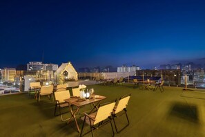 Un área de comedor al aire libre con mesas y sillas, una estructura parecida a una casa y vistas de la ciudad al anochecer.