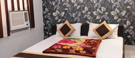 A modern hotel room with a large bed, a floral-patterned wall, a window with vertical blinds, and a built-in air conditioning unit.