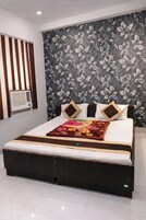 A modern hotel room with a large bed, a floral-patterned wall, a window with vertical blinds, and a built-in air conditioning unit.