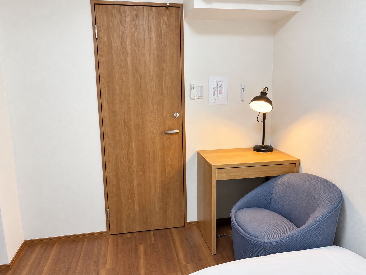 A small room with a wooden door, a desk with a lamp, and a blue chair.