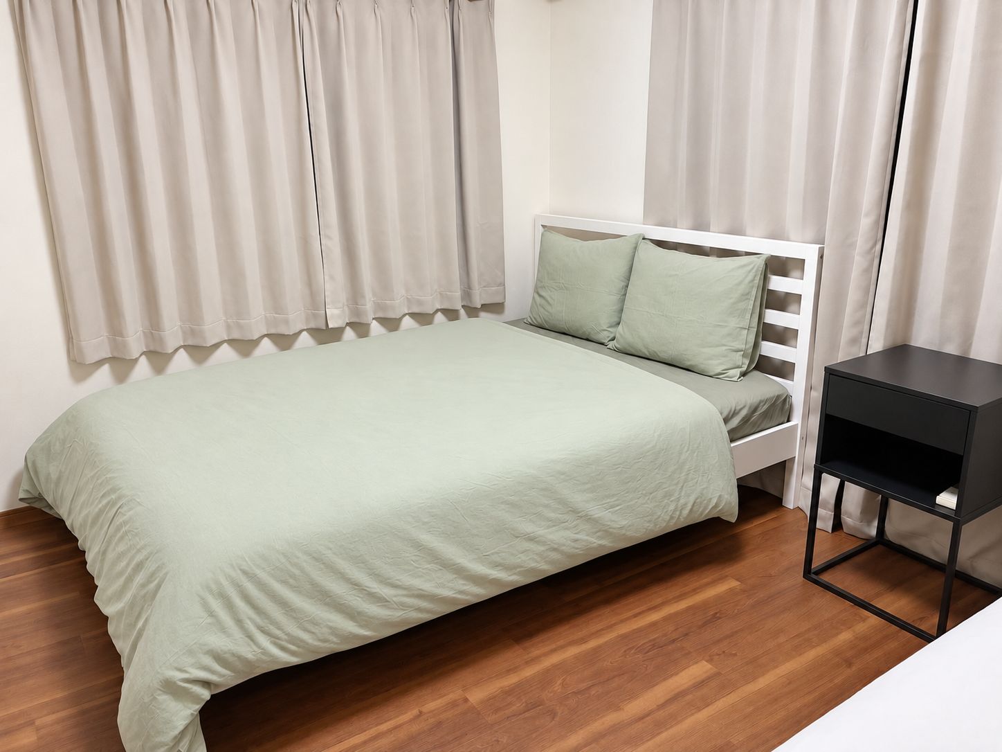 A single bed with a white frame and a green bedspread in a room with wooden flooring and beige curtains.