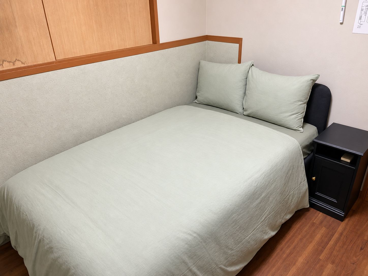 Een enkel bed met een houten hoofdeinde, een nachtkastje met lade en een lichtschakelaar op de muur.