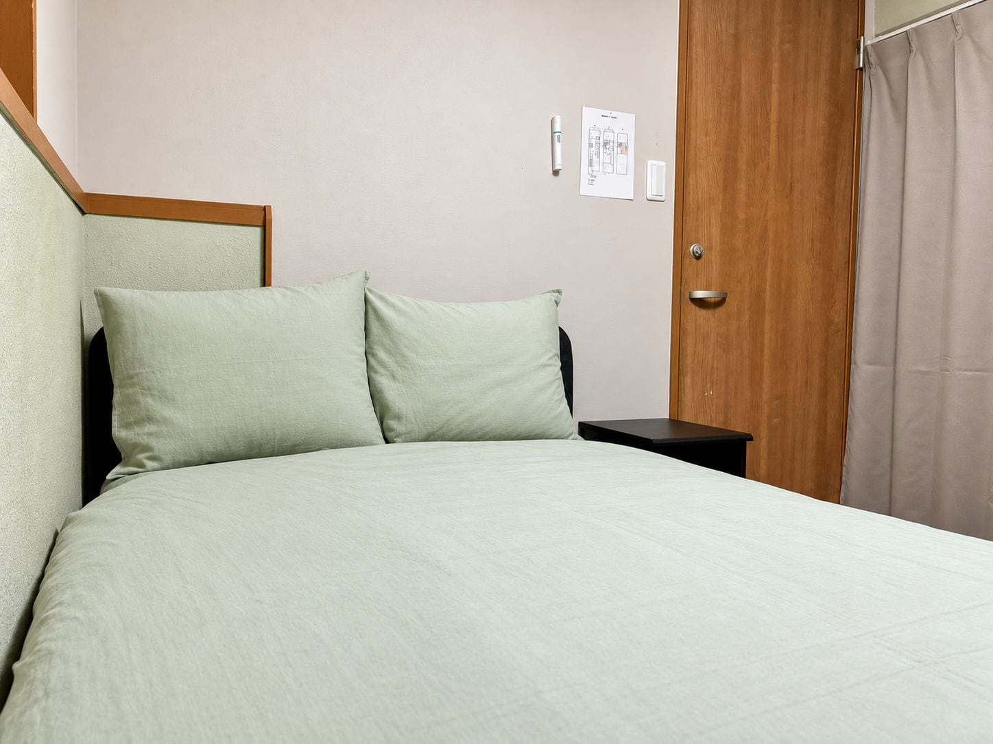 A single bed with a headboard, a nightstand, a door, and a wall-mounted light switch.