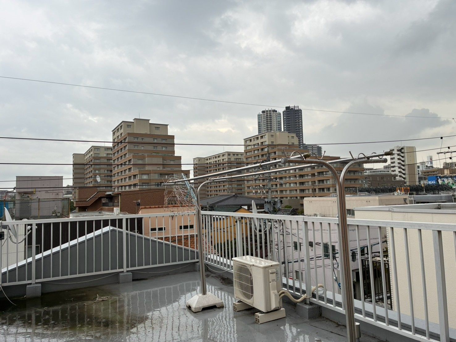 A rooftop terrace with a view of surrounding buildings, a metal railing, and an air conditioning unit.