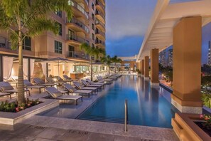 Un área junto a la piscina con sillones de descanso y una piscina, rodeada de palmeras y un edificio con balcones.