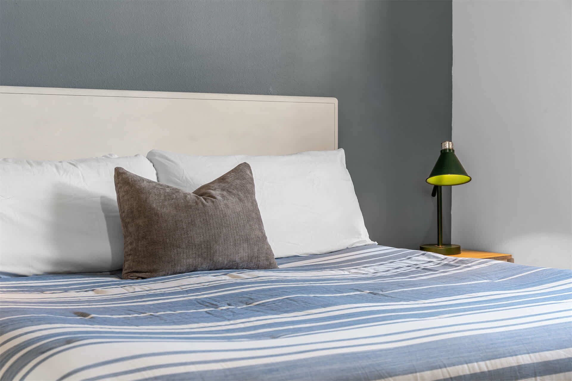 Una cama con colcha a rayas, una almohada y una mesita de noche con lámpara.