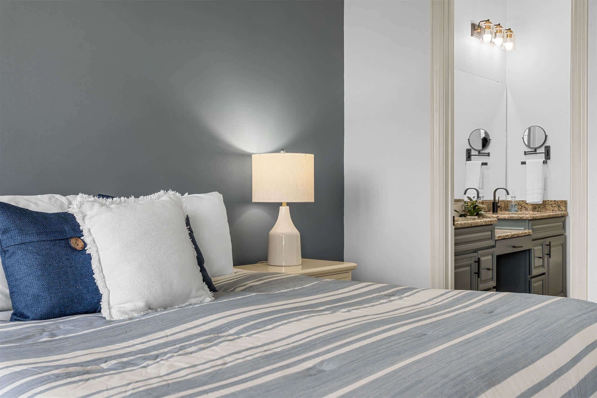 Un dormitorio con cama, lámpara de noche, mesita de noche, espejo y baño con lavamanos y encimera.