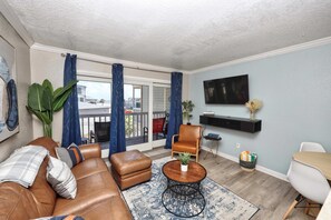 Ein Wohnzimmer mit braunem Ledersofa, einem Couchtisch, einem an der Wand befestigten Fernseher und einem Balkon mit blauen Vorhängen.