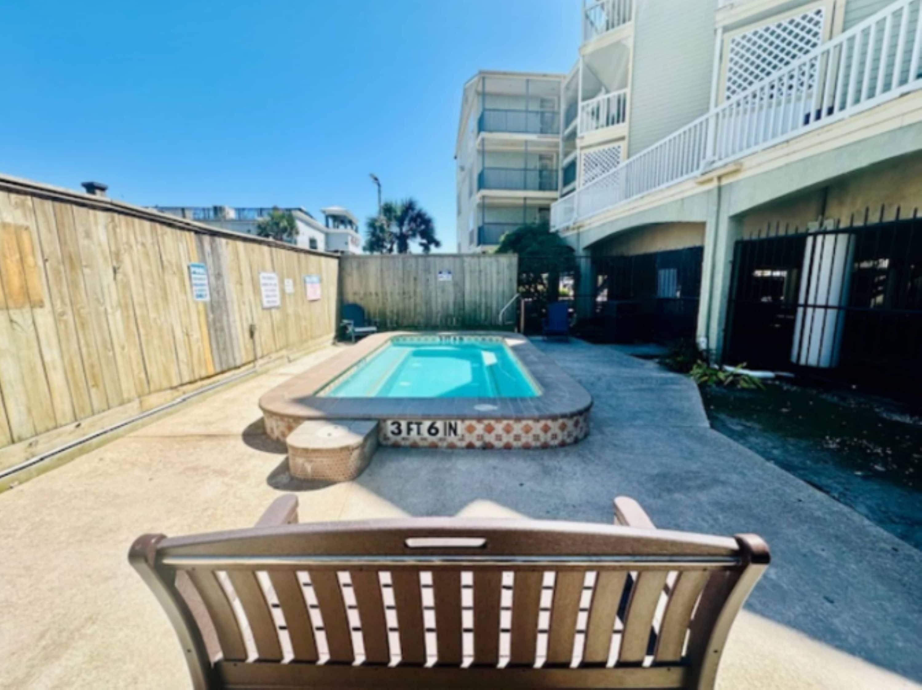 Un espace piscine entouré d’une clôture en bois, avec un banc et un bâtiment doté de balcons.