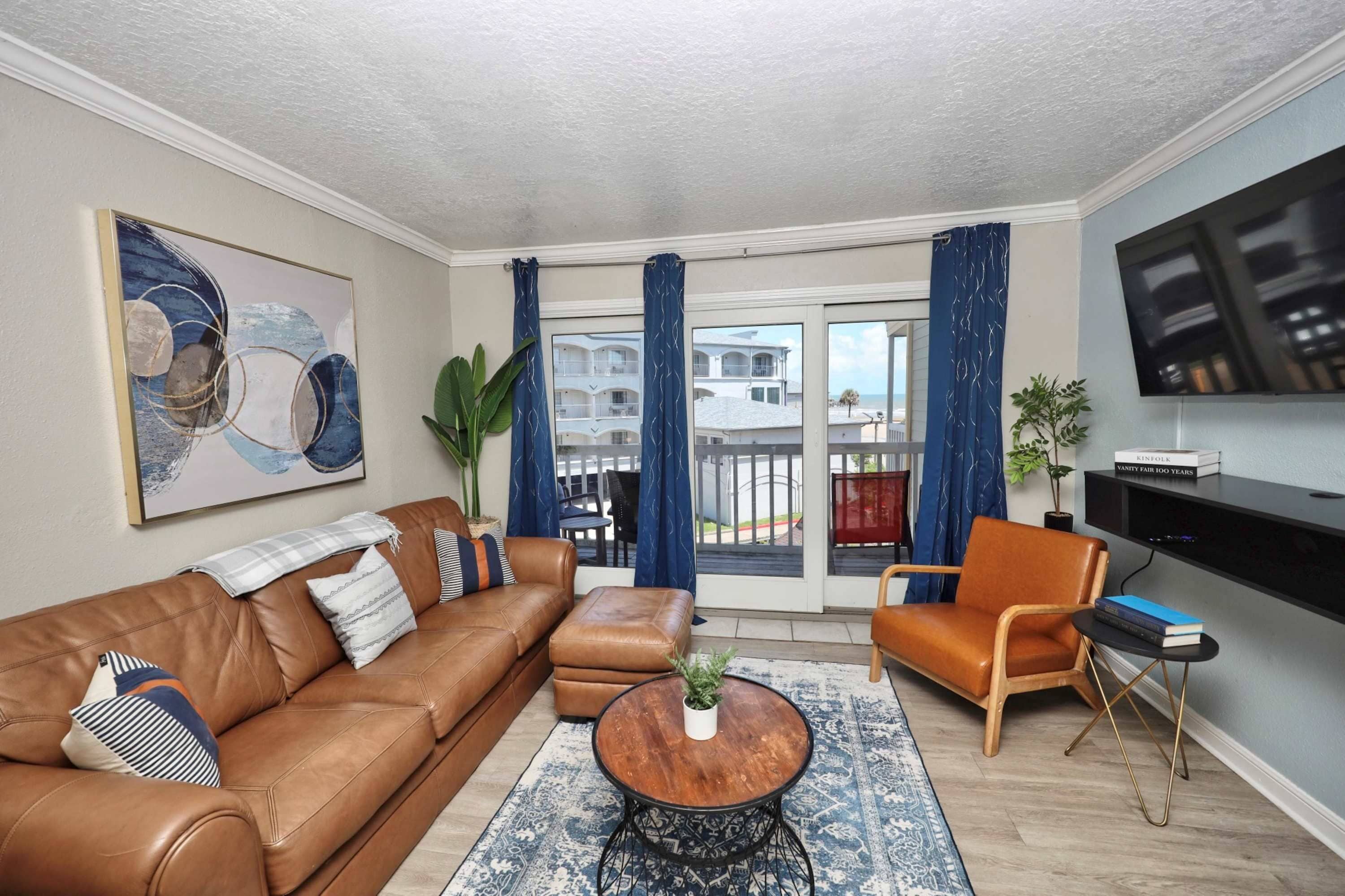 A living room with a brown leather sofa, a coffee table, a TV, and a view of a balcony with blue curtains.