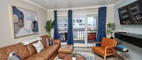 A living room with a brown leather sofa, a coffee table, a TV, and a view of a balcony with blue curtains.